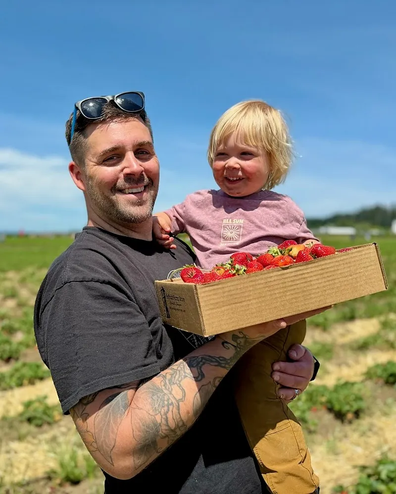 Brennan holding his young son Malakai and a box of fresh-picked strawberries in a sunny strawberry field